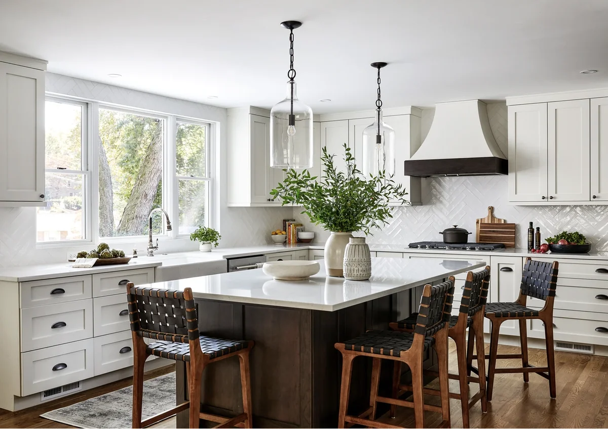 White kitchen with a dark island, woven chairs, a farmhouse sink and a herringbone tile backsplash.